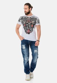 Weißes T-Shirt mit grafischem schwarz-rotem Design, kombiniert mit blauen, abgetragenen Jeans und beigen Sneakers. Das Outfit hat einen lässigen Schnitt.