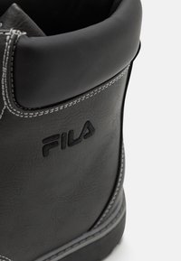 Botín negro de tobillo hecho de material sintético, con costuras blancas y un logo de Fila. Parte superior redondeada con una textura suave.