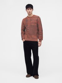 Homme portant un pull en maille orange et marron à motifs, un pantalon large noir et des chaussures marron, debout avec une main dans la poche.