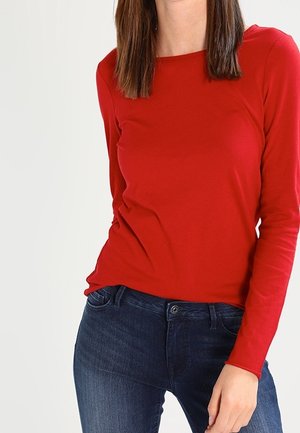 Vrouw draagt een strakke rode lange mouwen shirt en donkerblauwe spijkerbroek, staand tegen een effen lichte achtergrond.