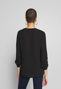Blouse noire à manches longues, en tissu léger, avec un col rond, des détails plissés sur les manches et un ourlet arrondi. Portée avec un jeans foncé.