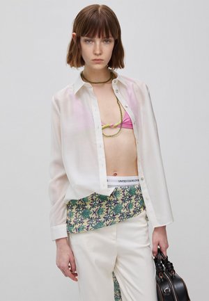 Junge Frau mit kurzen braunen Haaren trägt ein weißes transparentes Hemd über einem pinken Bikinioberteil, einen gemusterten Gürtel, weiße Hose und hält eine schwarze Tasche.