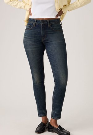 Nő sötétkék szűk farmernadrágban, fekete mokaszinban, fehér, haspólóval és sárga kardigánban, kezét csípőjén tartva.