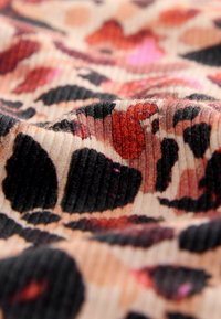 Tejido estampado con una textura ribeteada y manchas vibrantes en rojo, rosa y negro sobre un fondo beige. La vista en primer plano resalta los detalles.