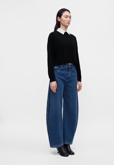 Femme debout portant un haut noir à manches longues avec col blanc, un jean bleu large et des bottines à talons noires sur un fond clair uni.