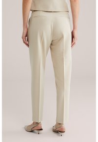 Vrouw draagt beige, op maat gemaakte broek en beige slingback schoenen met lage hak, staat ontspannen met handen langs het lichaam tegen een neutrale achtergrond.