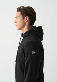 Sudadera negra con un bolsillo canguro en la parte delantera, capucha con cordón y un logo redondo de Colmar en la manga izquierda. Tejido suave, corte casual.