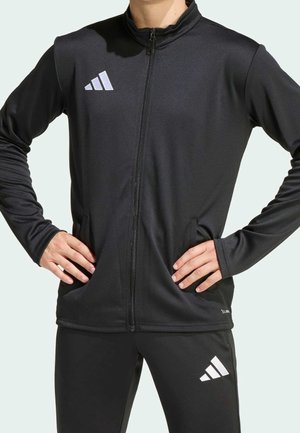 Osoba nosząca czarną kurtkę Adidas zapinaną na zamek błyskawiczny z rękami na biodrach, do kompletu czarne spodnie Adidas.
