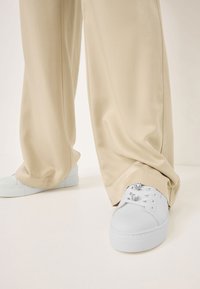 Sneaker bianchi in pelle con punta rotonda, suola piatta e dettagli metallici sui lacci. Abbinati a pantaloni beige larghi.