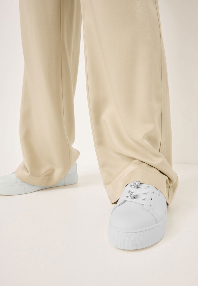 Sneaker bianchi in pelle con punta rotonda, suola piatta e dettagli metallici sui lacci. Abbinati a pantaloni beige larghi.