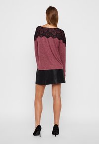 Bordeauxfarbenes Langarmoberteil mit schwarzem Spitzenüberzug an den Schultern, kombiniert mit einem schwarzen Leder-Minikleid und schwarzen High Heels.