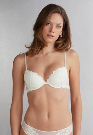Weißes Spitzen-Bralette mit gepolsterten Cups, verstellbaren Trägern und gewellten Kanten. Transparenter Mesh-Band unterhalb der Cups. Einfaches und zartes Design.