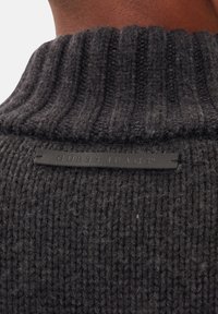 Dunkelgrauer, gestrickter Pullover mit einem gerippten Kragen; hinten mit einem schwarzen Lederetikett mit der Aufschrift "GUESS JEANS" versehen.