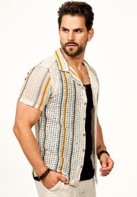 Kurze Ärmel, offen gestricktes Shirt in Creme mit mehrfarbigen Streifen (Orange, Blau, Grau) und Holzknöpfen. Über einem schwarzen Tanktop getragen.