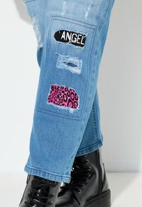 Světle modré džíny s oprýskaným vzhledem a nášivkami; "ANDĚL" v černé barvě a růžový leopardí vzor. Černé šněrovací boty doplňují outfit.
