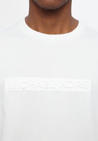 White cotton t-shirt with a round neckline and textured, raised "MICHAEL KORS" logo across the chest. Soft fabric, casual design.