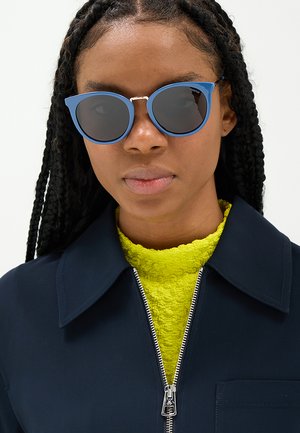 Donna con capelli intrecciati indossa occhiali da sole rotondi blu, una maglia gialla vivace e una giacca scura con cerniera argento.