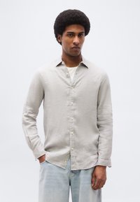 Νέος άνδρας με αφροαμερικάνικο χτένισμα, φορώντας ανοιχτού γκρι πουκάμισο πάνω από λευκό τοπ και ανοιχτό μπλε τζιν, στέκεται με το ένα χέρι στην τσέπη.