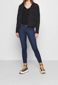 Chaqueta de cuadros en azul marino con cuello de borreguito, top negro de cuello en V, jeans ajustados azul oscuro y zapatillas altas negras con detalles en naranja.