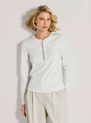 Blonde vrouw met kort golvend haar, gekleed in een witte lange mouwen top met decoratieve knopen en beige geplooide broek, staand tegen een lichte muur.