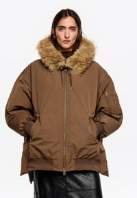 Chaqueta acolchada marrón con capucha forrada de piel, cremallera delantera y puños elásticos. Incluye bolsillos laterales y un diseño distintivo y estructurado.