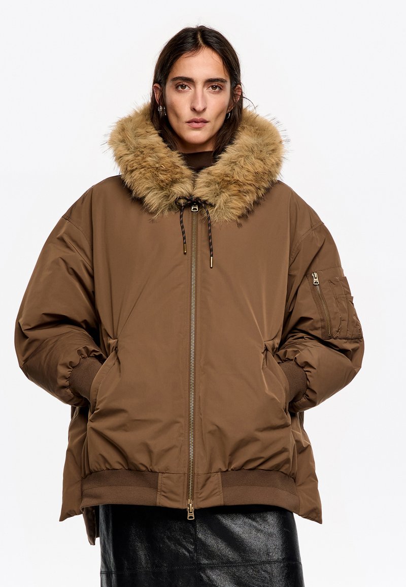 Chaqueta acolchada marrón con capucha forrada de piel, cremallera delantera y puños elásticos. Incluye bolsillos laterales y un diseño distintivo y estructurado.