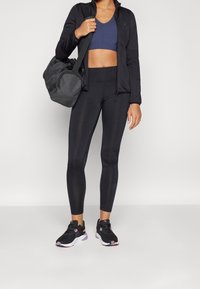 Chaqueta deportiva negra con cremallera, sujetador deportivo azul marino, leggings negros y zapatillas negras con acentos rosas. Texturas suaves, diseño ajustado.