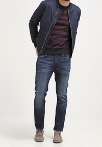 Mann trägt eine marineblaue Bomberjacke, dunkelroten und schwarzen gestreiften Pullover, blaue Jeans und hellbraune Schnürstiefel, steht mit den Händen hinter dem Rücken.