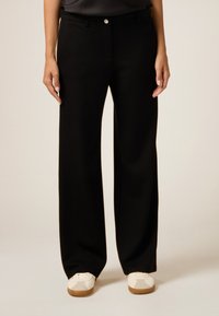 Pantalons larges noirs avec une texture lisse, dotés d'une fermeture par bouton à l'avant et d'une ceinture, associés à des baskets blanches sur un fond clair.