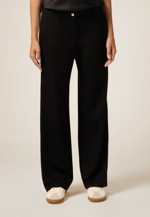 Pantalons larges noirs avec une texture lisse, dotés d'une fermeture par bouton à l'avant et d'une ceinture, associés à des baskets blanches sur un fond clair.