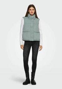Giovane donna che indossa un gilet imbottito grigio chiaro sopra una maglietta bianca a maniche lunghe, jeans skinny neri e stivaletti neri, in piedi su uno sfondo neutro.