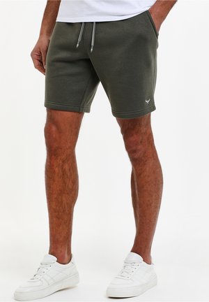 Mand iført mørkegrønne shorts med snor og lommer samt hvide sneakers, stående mod en ensfarvet lys baggrund.