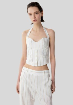 Donna che indossa un top all'americana bianco a righe sottili con bottoni e pantaloni abbinati a vita alta, in piedi davanti a uno sfondo grigio.