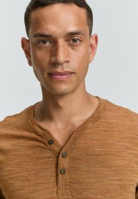 Bruine Henley-shirt met een textuurstof, voorzien van een V-hals en twee donkere knopen op de knoopsluiting. Eenvoudig ontwerp met subtiele kleurvariaties.