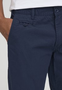 Personne portant un pantalon bleu marine avec une petite poche boutonnée au-dessus de la main droite, positionnée près de la ceinture.