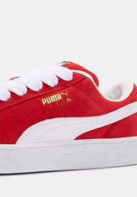 Red suede sneaker with white rubber sole, featuring white laces, gold branding, and a green accent on the heel. Classic design.