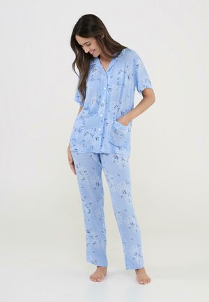 Mujer con un conjunto de pijama floral azul claro, con camisa de manga corta y botones y pantalones a juego, de pie descalza sobre un fondo blanco.