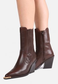 Piernas que llevan botas de cuero marrón oscuro hasta la mitad de la pantorrilla, con puntera estilizada, tacones de bloque y cremallera lateral, sobre un fondo blanco.