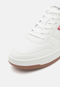 Weiße Leder-Sneaker mit perforiertem Design, weißen Schnürsenkeln und einer braunen Gummisohle. Mit einem roten Logodetail an der Seite.