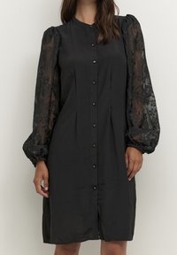 Black knee-length button-down dress with sheer puffed lace sleeves featuring floral embroidery, worn by a standing person against a plain background.