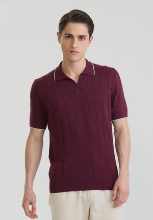 Młody mężczyzna w bordowej koszulce polo z krótkim rękawem i białym wykończeniem oraz jasnobeżowych spodniach, stojący na jednolitym tle.
