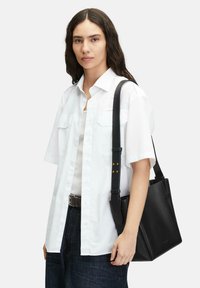 Czarna skórzana torba na ramię ze sztywnymi bokami i regulowanym paskiem, w zestawieniu z białą koszulą z krótkim rękawem na jasnej bluzce na ramiączkach.
