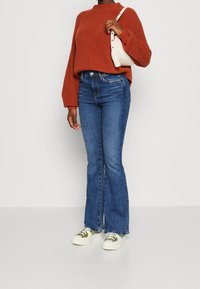Rostfärgad ribbad tröja, blå utfällda jeans och krämfärgade sneakers med gröna detaljer. Modellen håller en vit handväska.