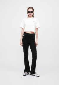Femeie îmbrăcată în pantaloni negri evazați, tricou alb scurt, pantofi plata argintii și ochelari de soare negri, stand în fața unui fundal simplu de culoare deschisă.