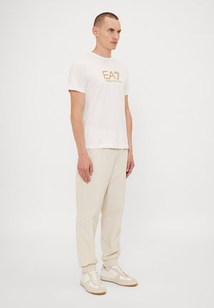 Camiseta blanca de algodón con el logo dorado "EA7", combinada con joggers taper de color beige claro y zapatillas blancas con acentos texturizados y suelas de goma.