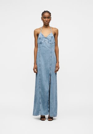 Γυναίκα με πλεξούδες στα μαλλιά, που φορά ένα αμάνικο μακρύ φόρεμα από ανοιχτό μπλε τζιν με λουριά γύρω από το λαιμό και σκίσιμο μπροστά, στέκεται μπροστά σε ουδέτερο φόντο.