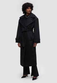 Černý dlouhý kabát vyrobený z vlněné směsi, s širokým límcem, páskem v oblasti pasu, velkými manžetami a bočními kapsami. Doplněný o černé úzké kalhoty.