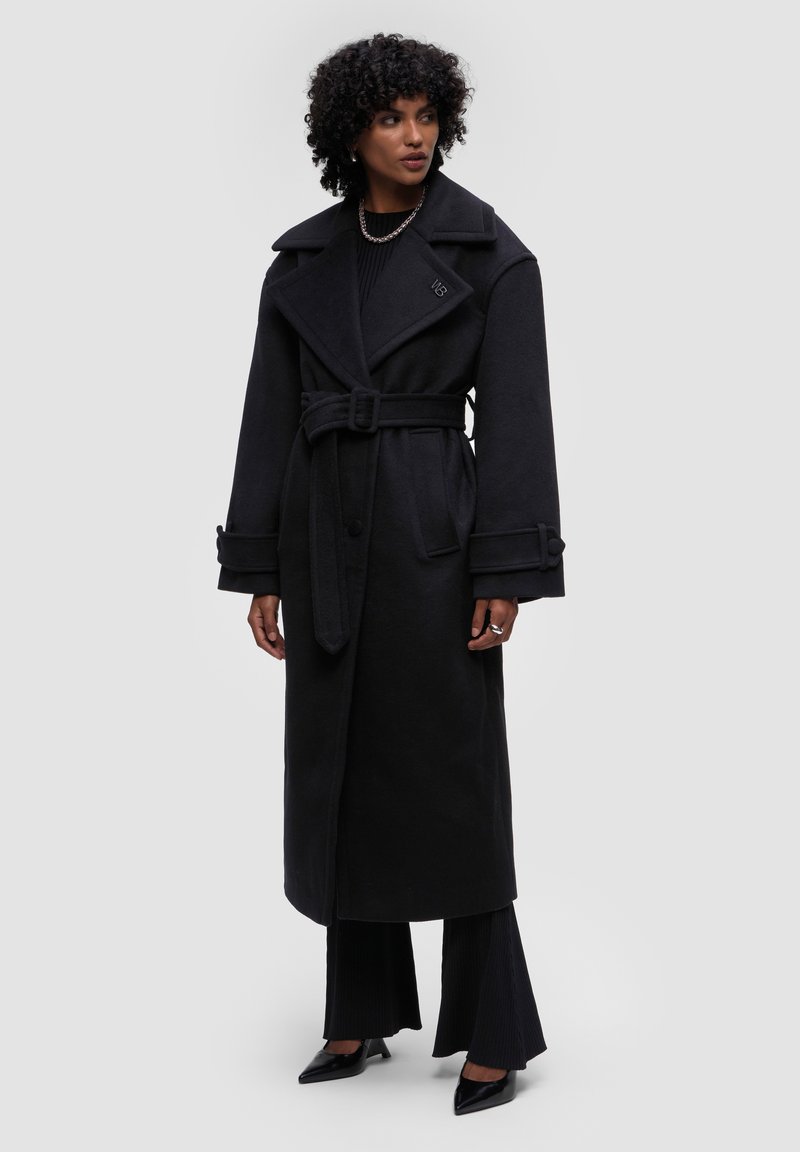 Černý dlouhý kabát vyrobený z vlněné směsi, s širokým límcem, páskem v oblasti pasu, velkými manžetami a bočními kapsami. Doplněný o černé úzké kalhoty.