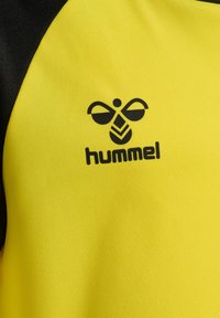 Gul sportstrøje med sorte accenter, der har en sort "hummel"-logo og et bi-design. Materialet er glat og tekstureret.