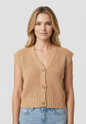 Donna bionda che indossa un gilet beige lavorato a maglia senza maniche con bottoni e jeans azzurri, in piedi davanti a uno sfondo grigio semplice.